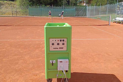 Grüne Ballmaschine steht im Vordergrund, hinter dem Tennisnetz warten zwei Tennispielerinnen Tennisunterricht in Philippsreut in Bayern